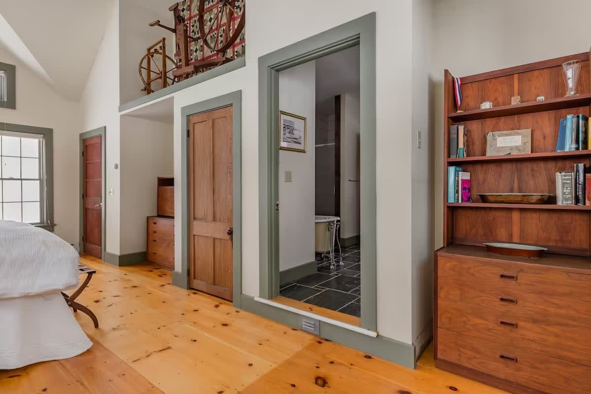 View of a bedroom with wooden floors, a bed partially visible on the left, a door to an adjacent room on the right, and a wooden bookshelf with books and decorative items on the far right. A small bathroom with dark tiled floor is visible through an 