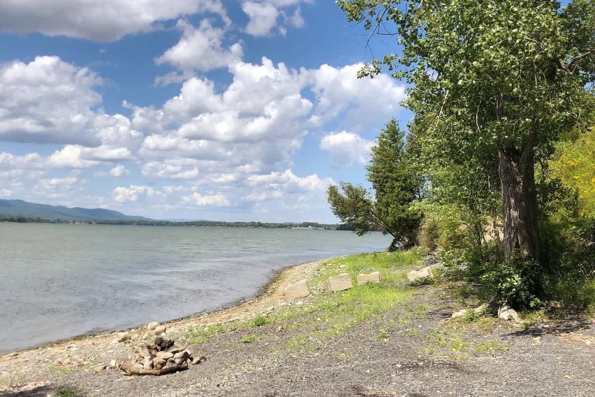A lakeside scene with a rocky shoreline, green grass, and trees on the right, and a wide lake under a partly cloudy sky.