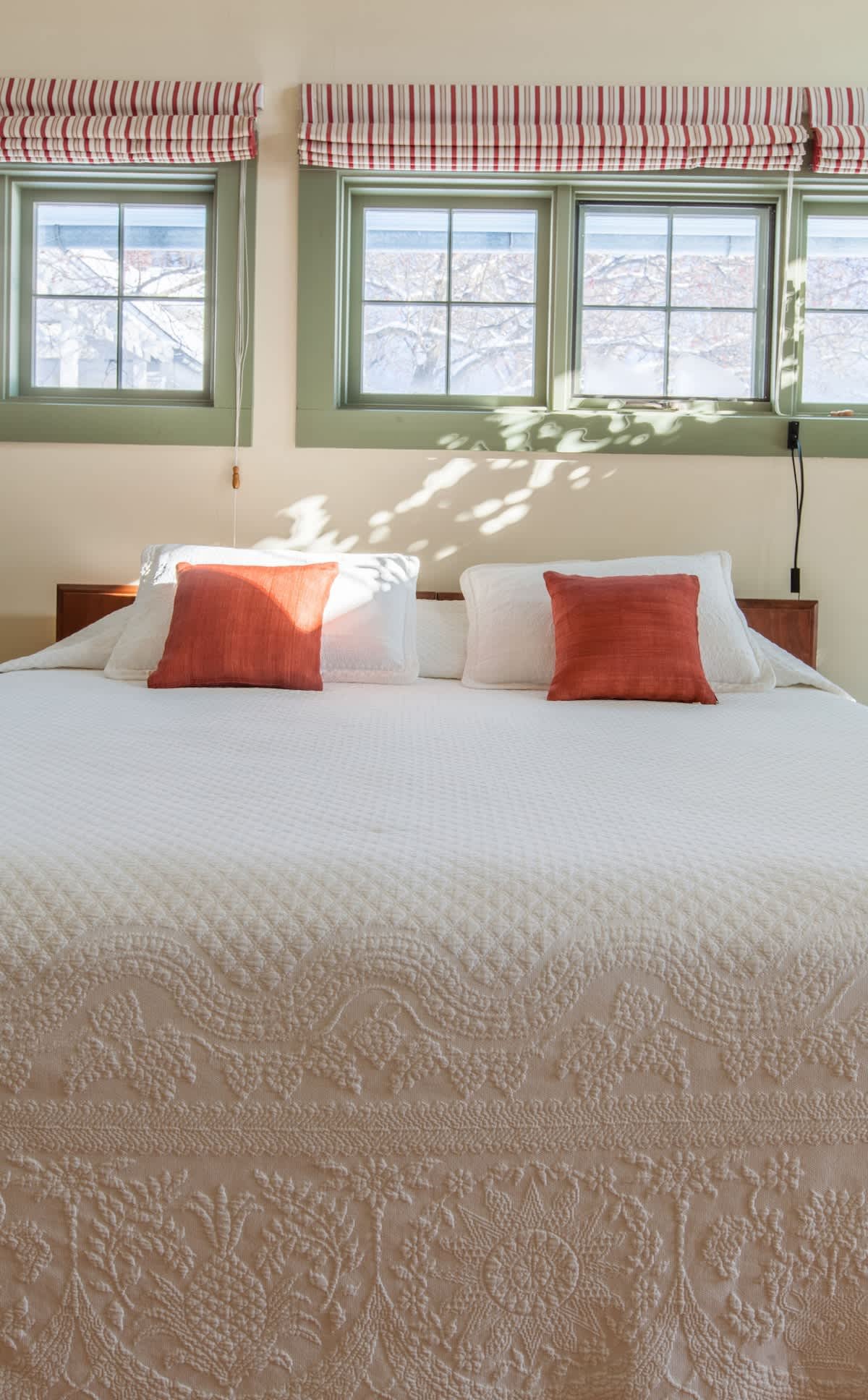 A neatly made bed with white textured bedding, three pillows with white pillowcases and two smaller rust-colored accent pillows, in front of a wall with three green-trimmed windows and striped valances, sunlight casting shadows on the bedding.