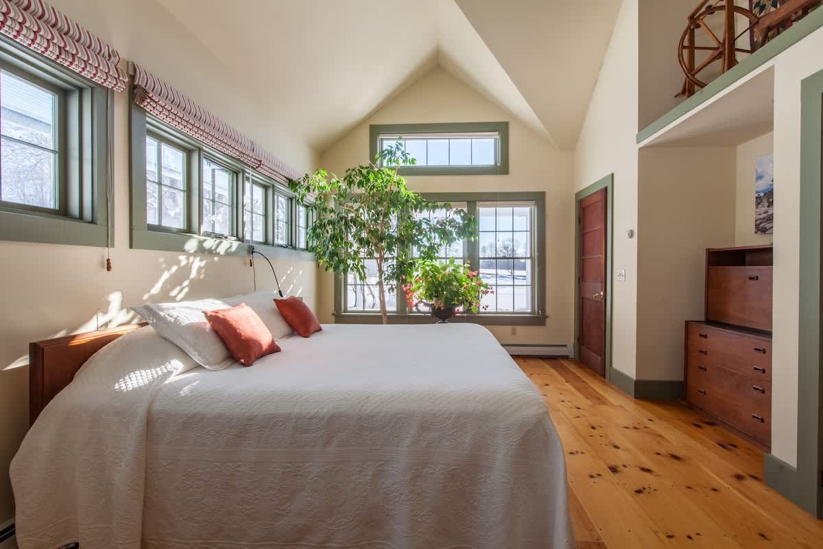 A cozy bedroom with a large bed covered in white bedding and pillows. There are multiple windows letting in natural light, with green trim on the windows. A tall plant is beside the window, and a wooden dresser is on the right side of the room. The c