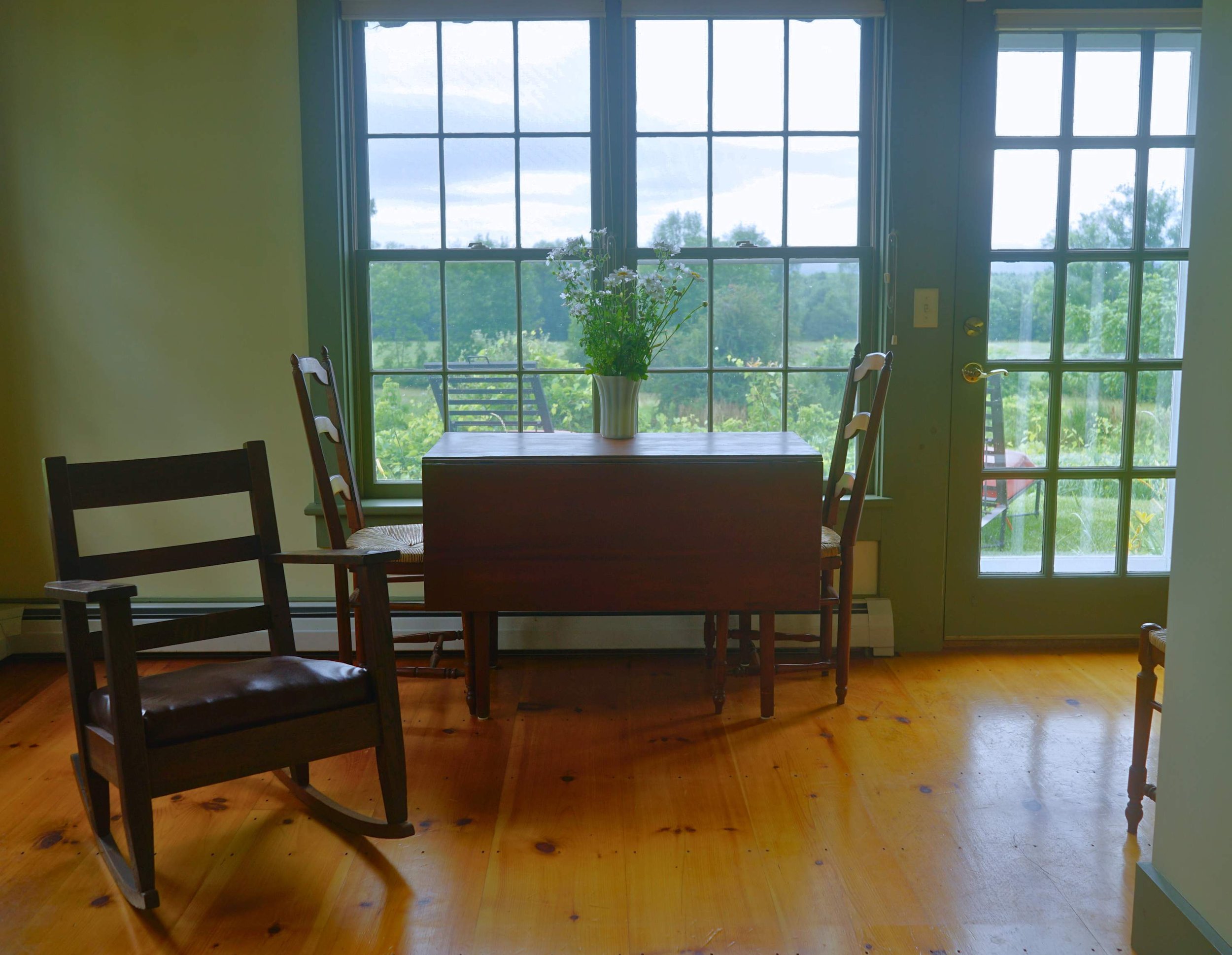 A wooden table with a vase of white flowers, two chairs by the window, a rocking chair, and a glass door leading outside, with a green landscape visible through the windows.