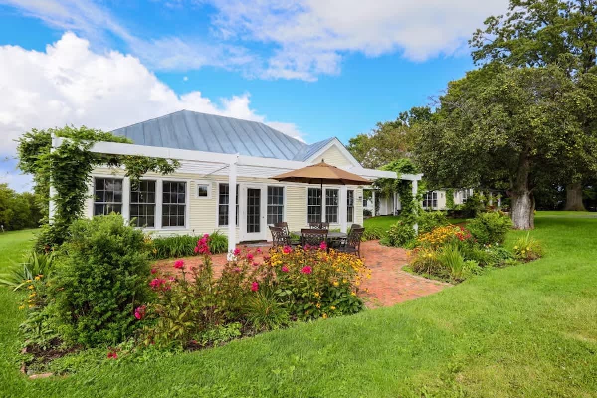 Serene guest house with white trim, surrounded by a lush green lawn and colorful garden beds with flowers, on a bright day with blue sky and clouds.