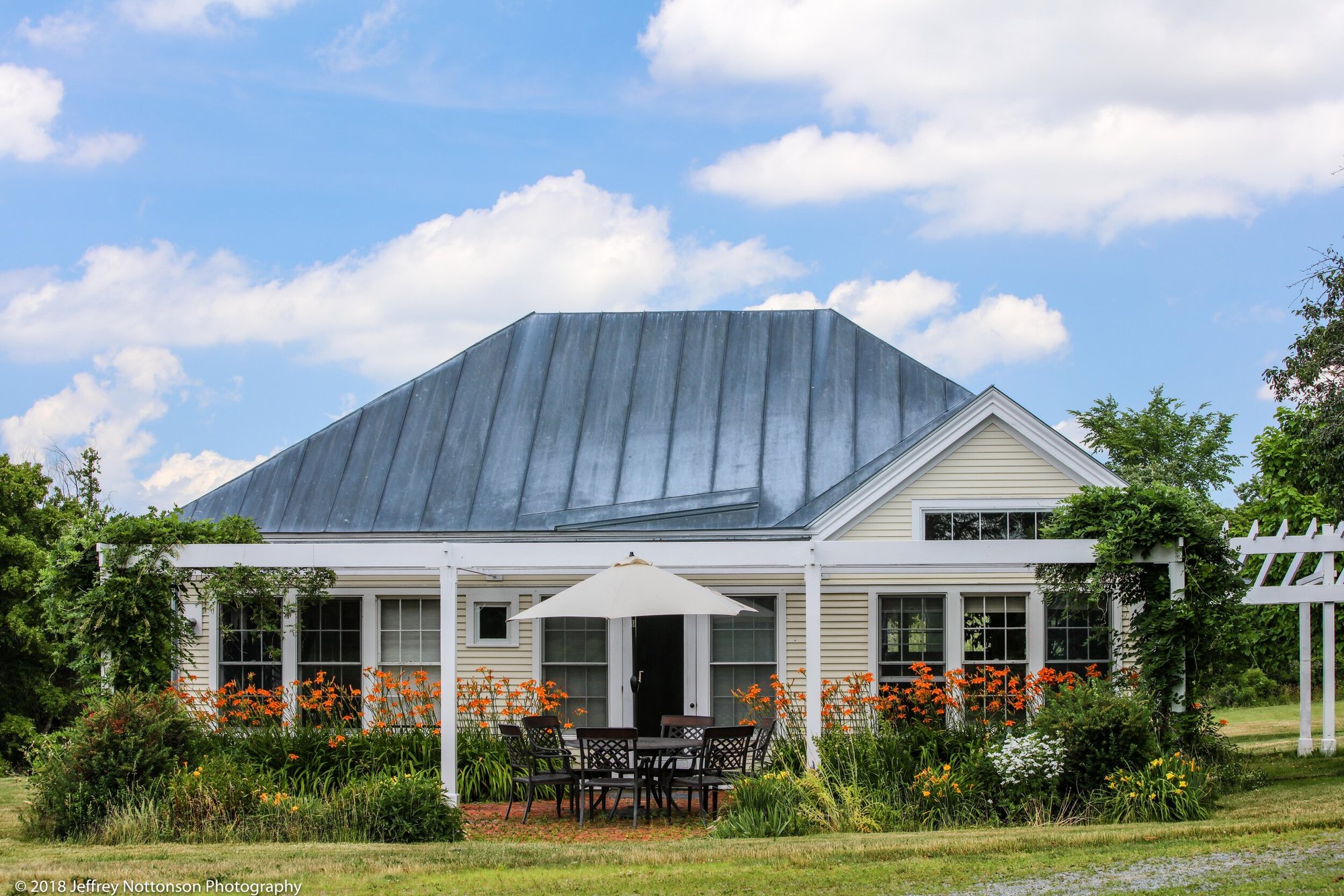 Guest house with standing-seam metal roof and white patio umbrella, framed by orange tiger lilies and perennial gardens.