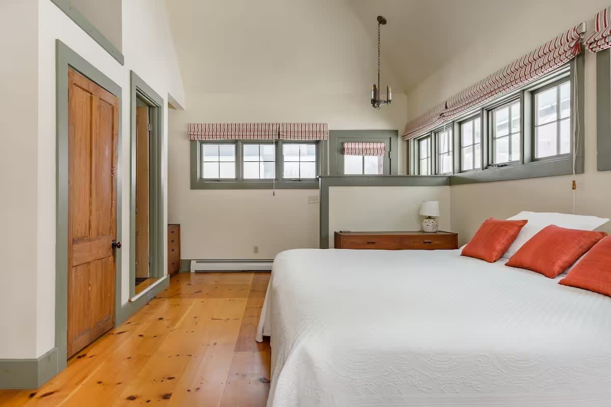 Bedroom with white bed, three orange pillows, wooden nightstand, lamp, multiple windows with plaid valances, wooden door, light-colored walls, wood floors, and a hanging light fixture.