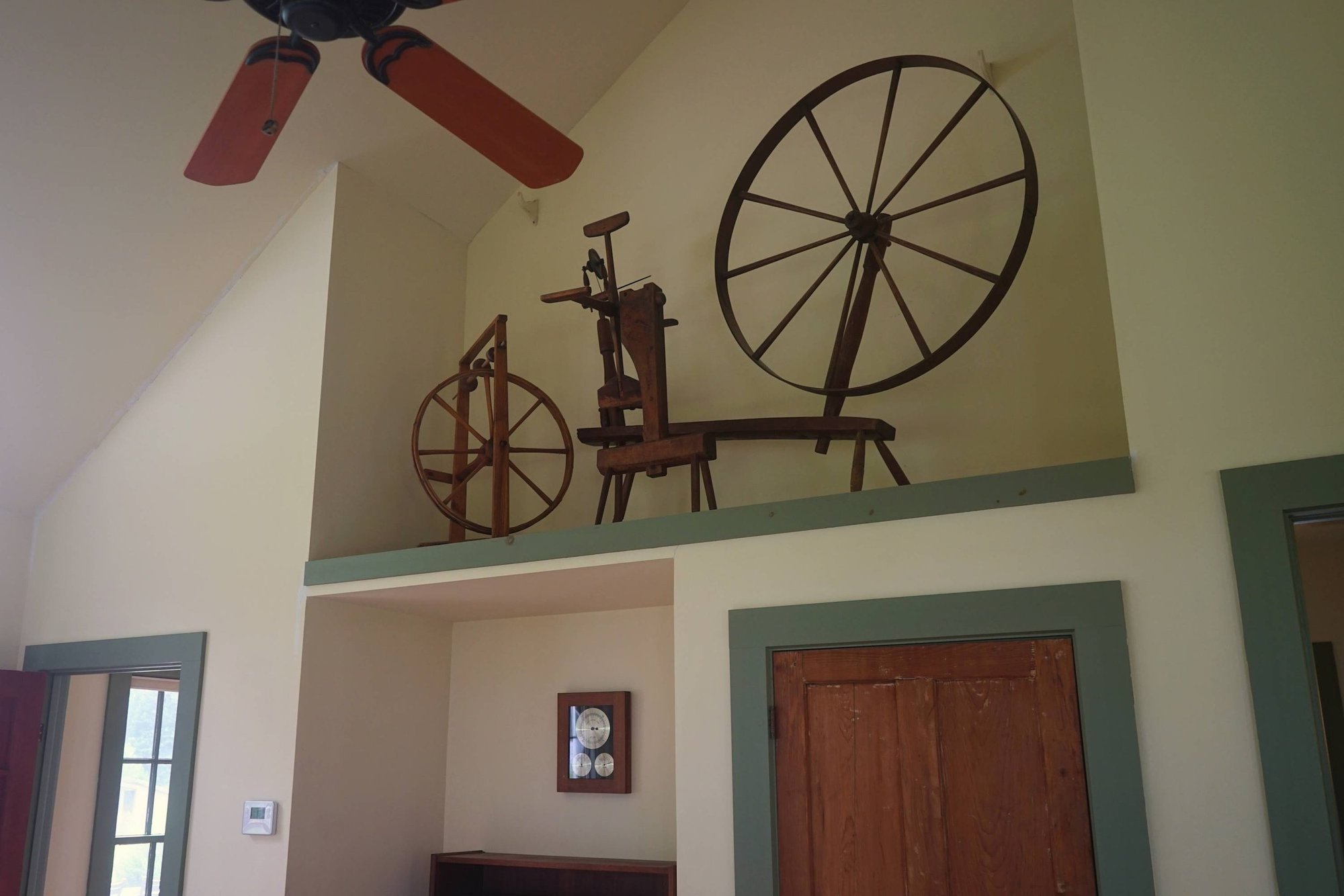 Decorative wooden wheels and spinning wheel on a ledge near ceiling in a room with green door and window frames.