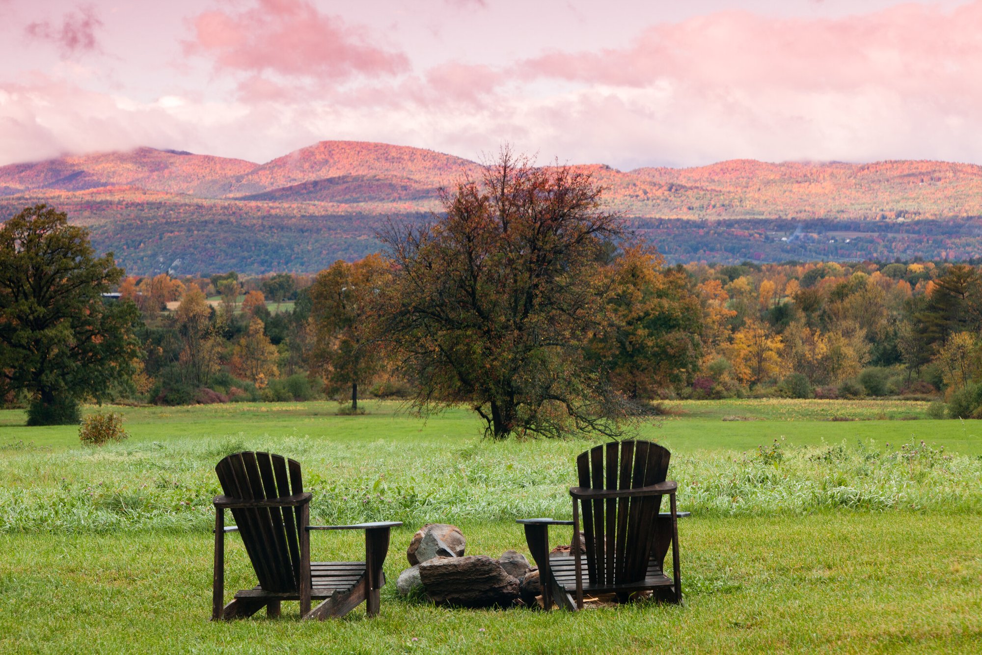 Two Adirondack chairs beside a fire pit in the meadow at sunset, with pink light over the Adirondack Mountains.